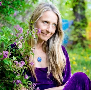 A woman with long blonde hair in a purple outfit sitting outdoors among purple flowers and green foliage.