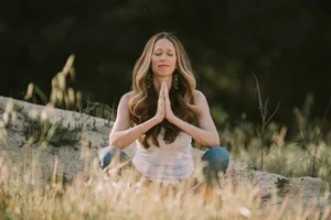 A woman practicing yoga outdoors, sitting on grass with her hands in prayer position.