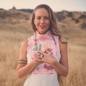 A woman standing outdoors in a field with dry grass, wearing a white dress and a pink embroidered scarf, smiling and holding her hands over her chest.
