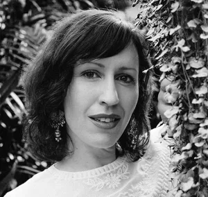 Black and white photo of a woman with shoulder-length dark hair, wearing earrings and a lace top, standing outdoors among trees and foliage.