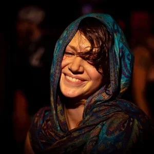 Smiling woman with short dark hair, wrapped in a colorful scarf, in a dimly lit environment.