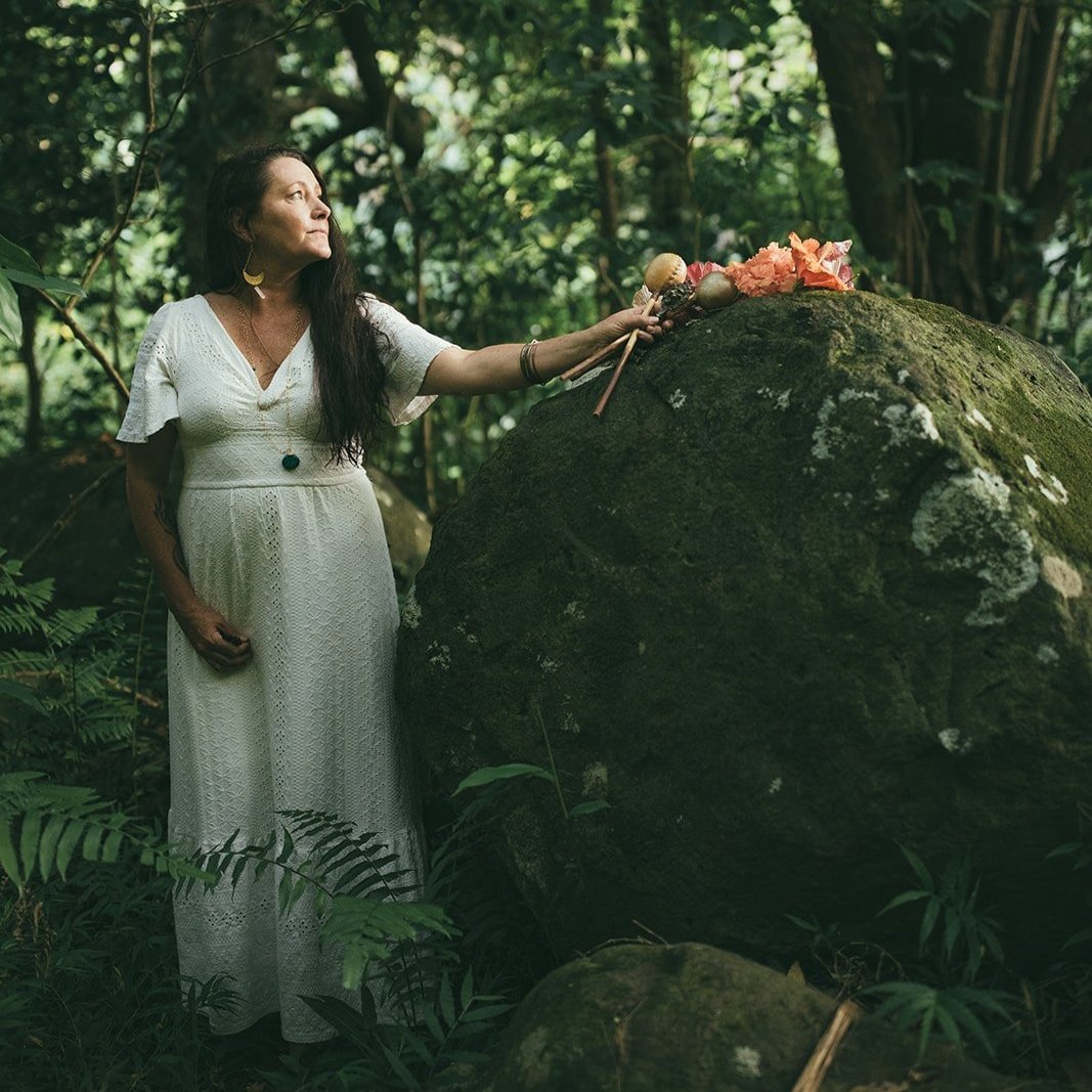 A woman in a white dress with long dark hair stands in a lush green forest next to a large moss-covered rock. She holds a small ceremonial stick and appears to be participating in a ritual, with flowers and objects placed on the rock.