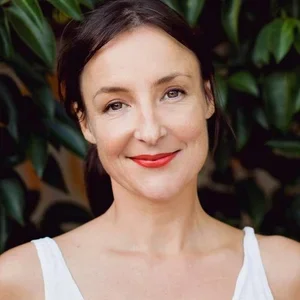 A woman with dark hair and red lipstick smiling, standing in front of green foliage.
