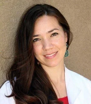 A woman with long dark hair smiling, wearing a white blazer and earrings, standing against a neutral background.