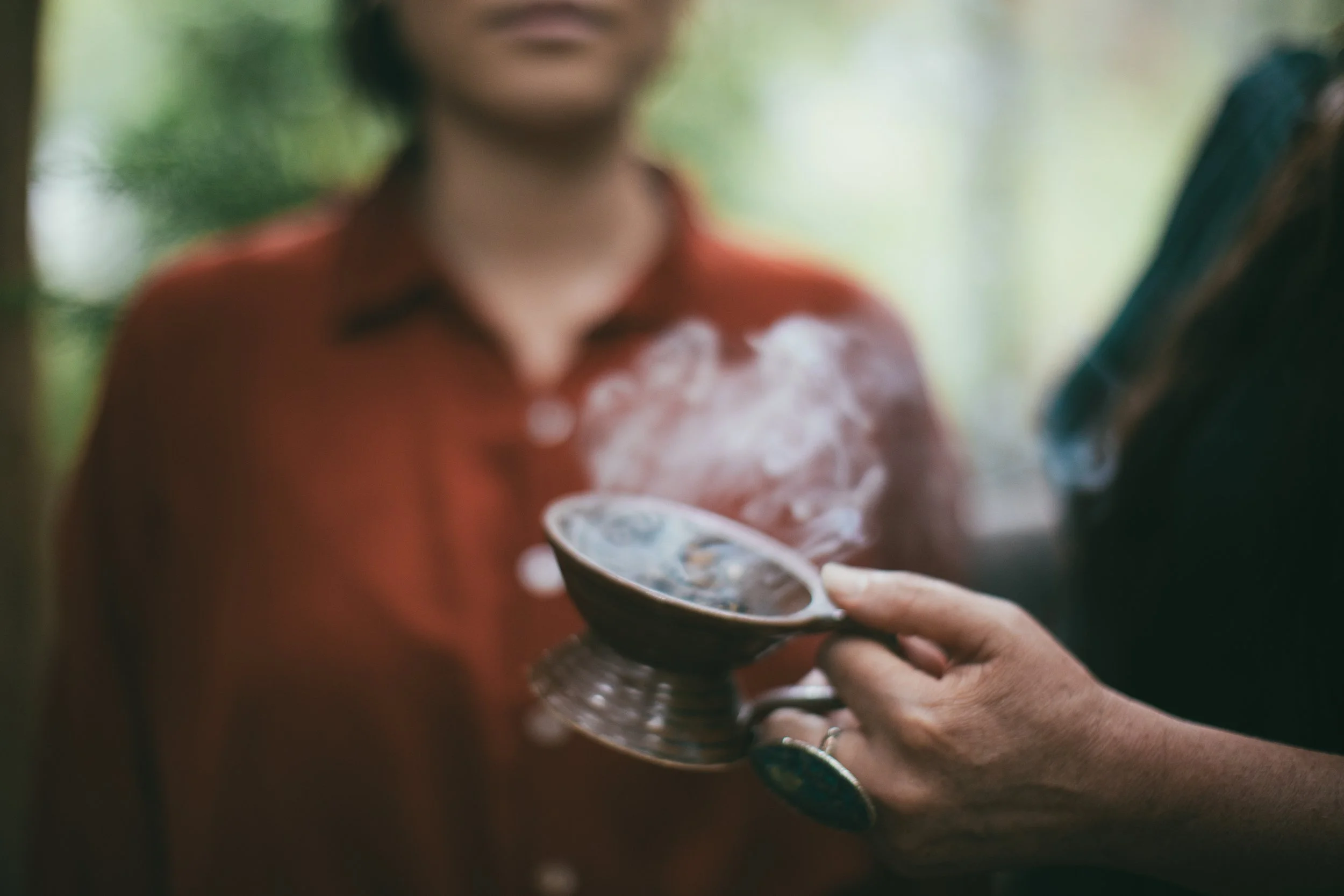 Person holding a small steaming bowl, with blurred woman in red shirt in background.