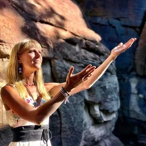 Young woman with blond hair wearing colorful jewelry and a light top, gesturing upward with open arms in a rocky outdoor setting.
