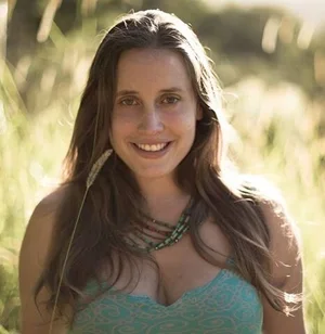 A woman with long brown hair smiling outdoors in sunlight, wearing a teal dress and layered necklaces.