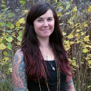 Young woman with dark hair and tattoos standing outdoors among tall grasses and yellow leaves.