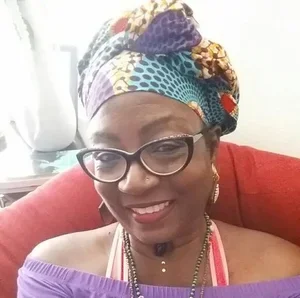 Woman wearing glasses, colorful headwrap, purple top, and jewelry, smiling in a cozy setting.