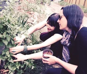 Two women gardening outdoors, one holding a glass, the other reaching for plants.