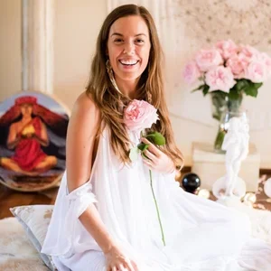 Smiling woman in a white dress holding a pink flower, sitting on a bed in a decorated room with pink flowers and art.