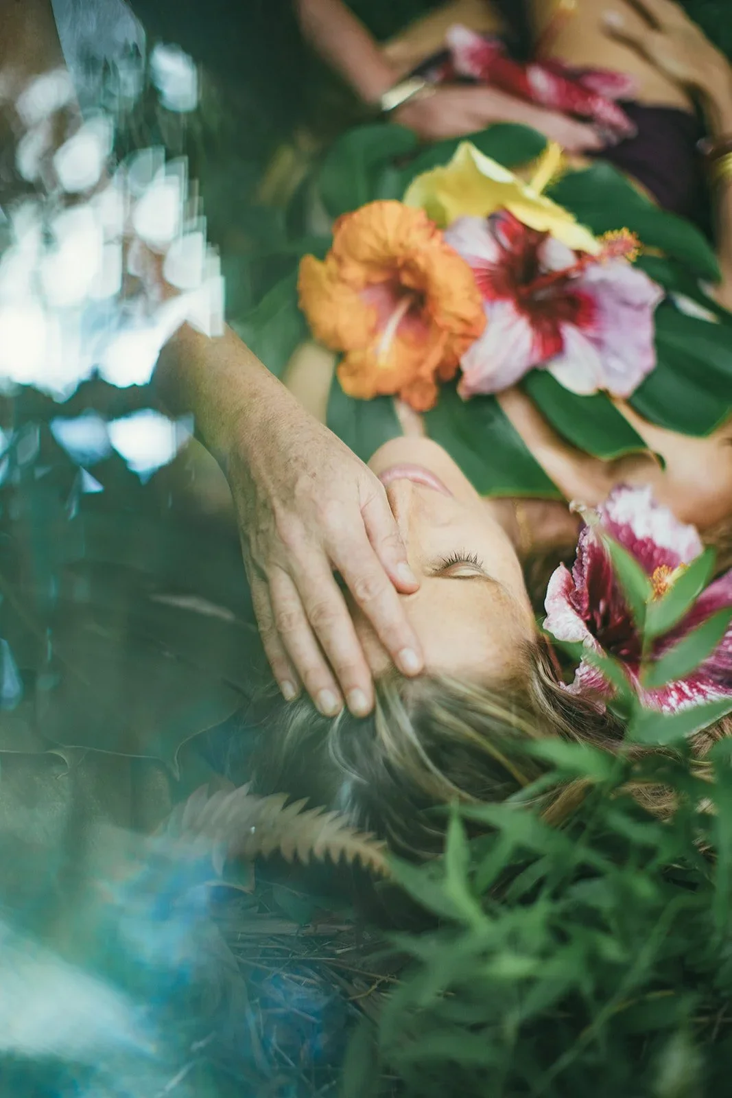 A woman lying on the ground surrounded by large pink and yellow flowers and green leaves, with a hand gently resting on her head.
