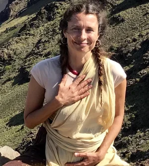 A woman in traditional clothing sitting outdoors on a rocky hillside with greenery in the background.