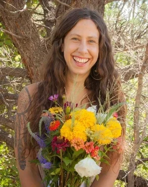 A woman with long wavy hair holding a colorful bouquet of flowers outdoors, smiling in front of a tree with green leaves.