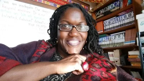 A woman with glasses and black curly hair smiling in a library, with shelves of books behind her.