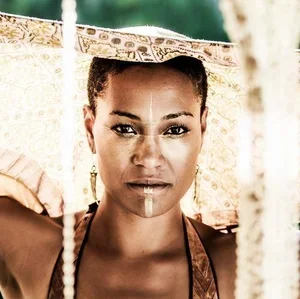 Close-up portrait of a woman with dark skin, braided hair, wearing traditional jewelry and clothing, outdoors with trees and a decorative umbrella in the background.