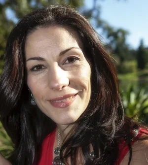 A woman with long dark hair smiling outdoors with trees and blue sky in the background.