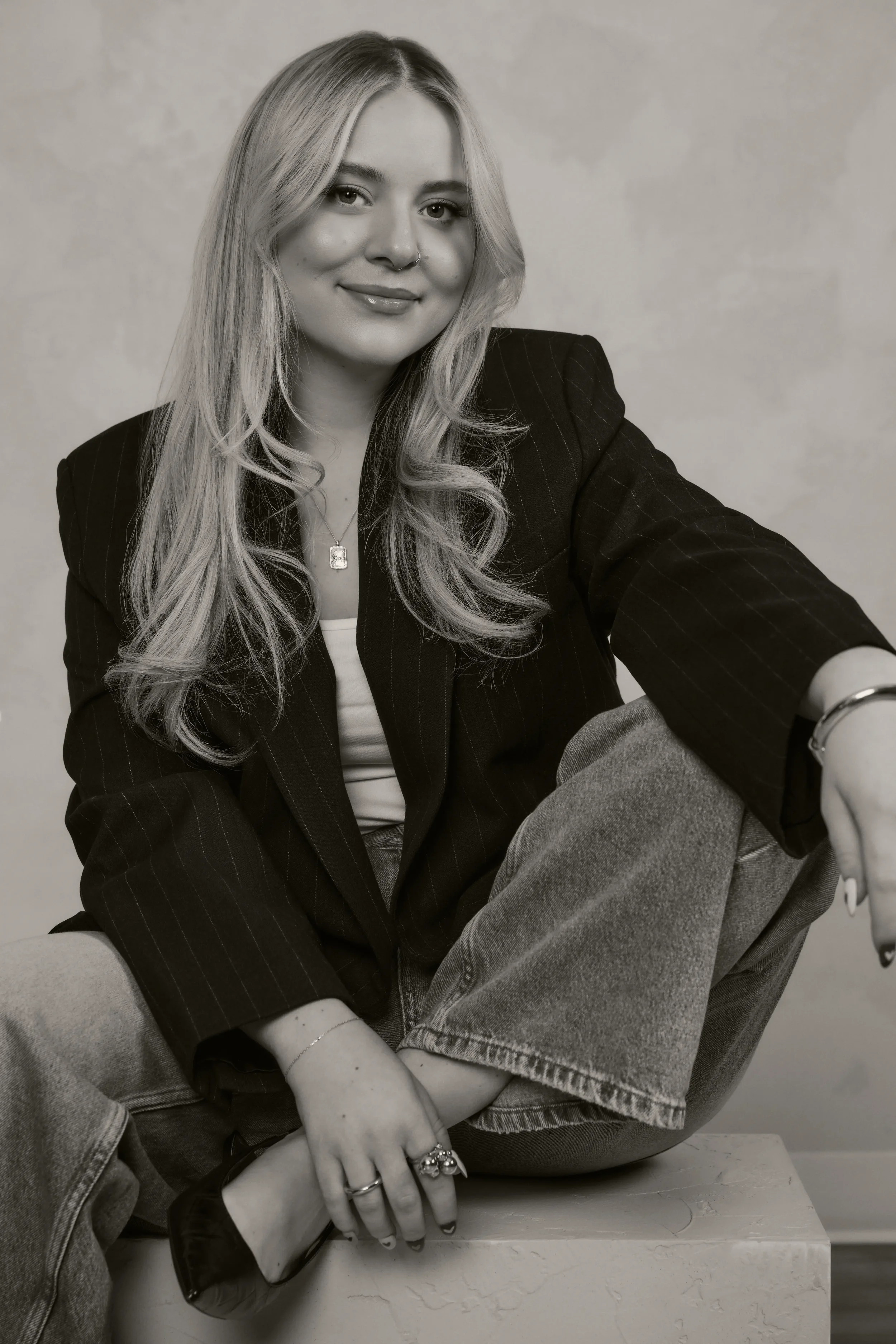 Portrait d'une femme assise, portant un blazer à rayures, avec des cheveux longs et blonds, souriant légèrement, avec des bijoux, dans un style monochrome.