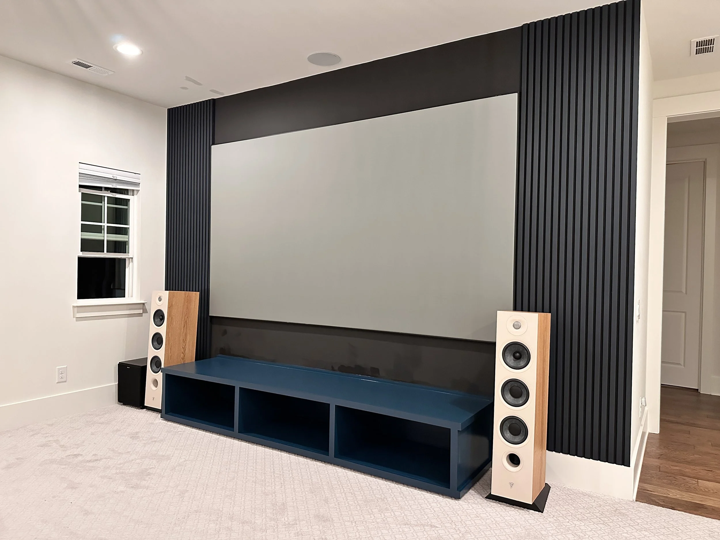 Home theater room with large gray screen, black acoustic panel, blue storage unit, two white floor-standing speakers, and a small black subwoofer.