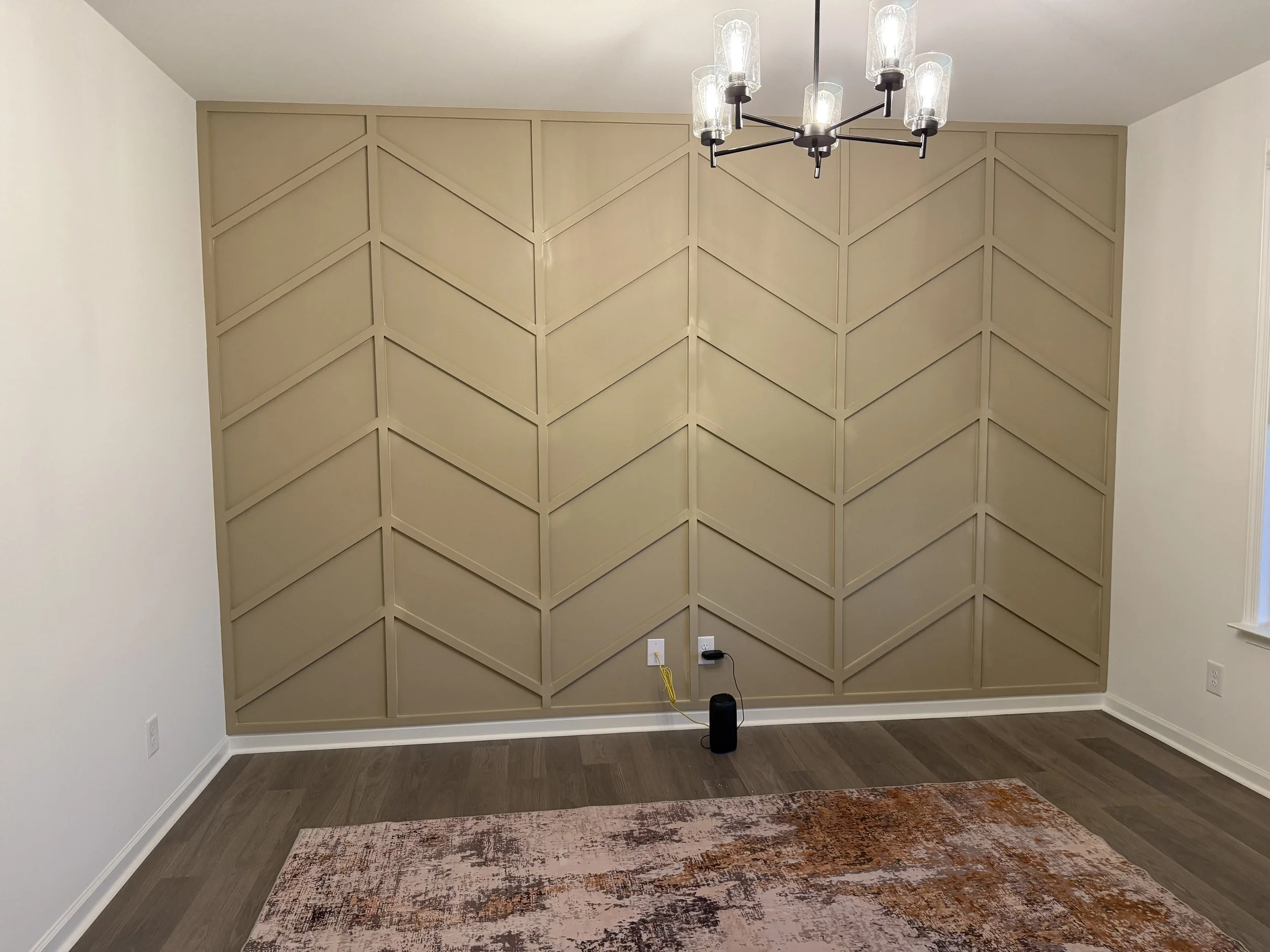 Empty room with a chevron-patterned beige accent wall, a chandelier, a colorful rug, and an outlet with cords.