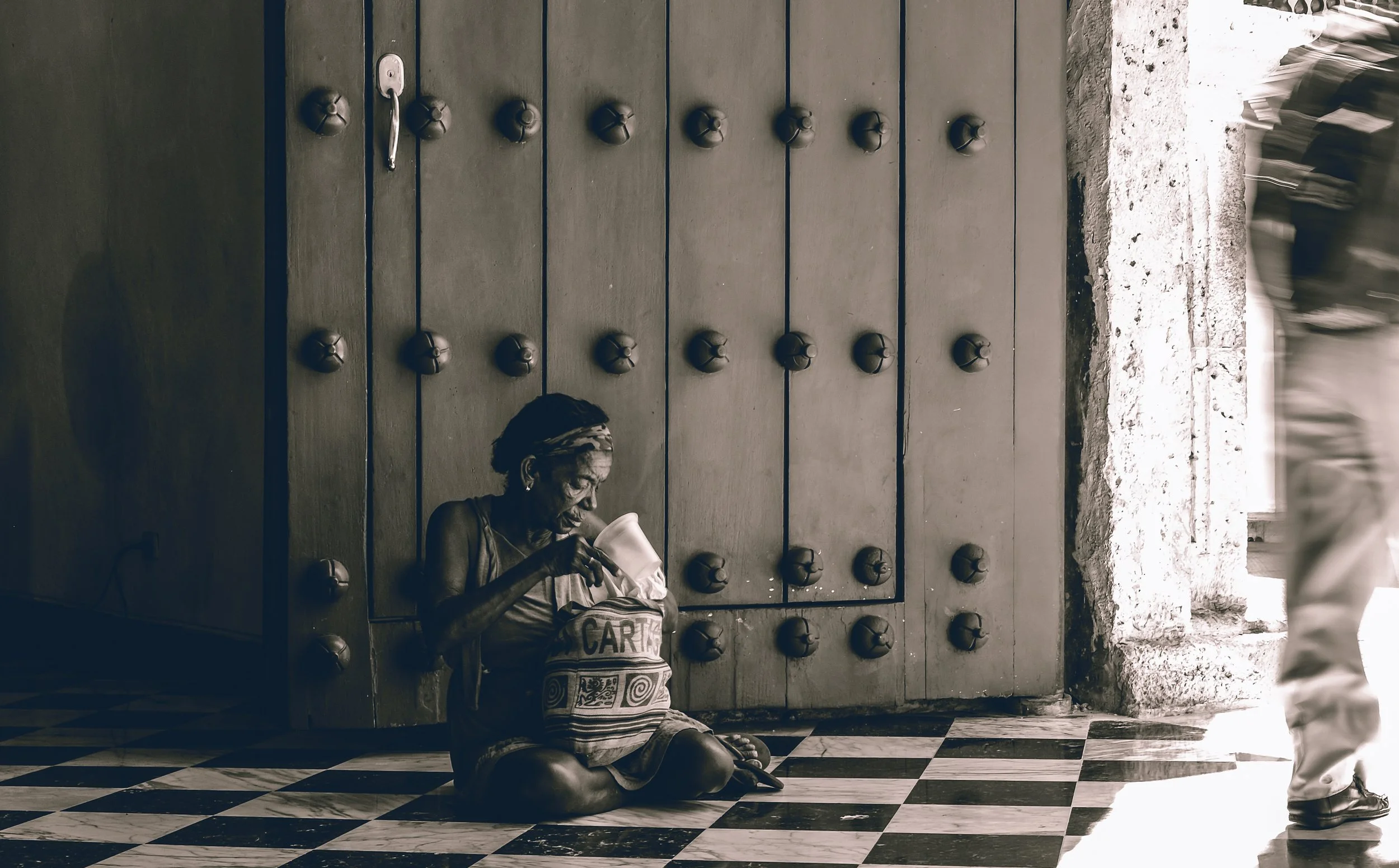 An elderly woman sitting on a checkered tile floor, drinking from a cup, leaning against a large wooden door with round metal knobs, with a person walking past in blurred motion to the right.