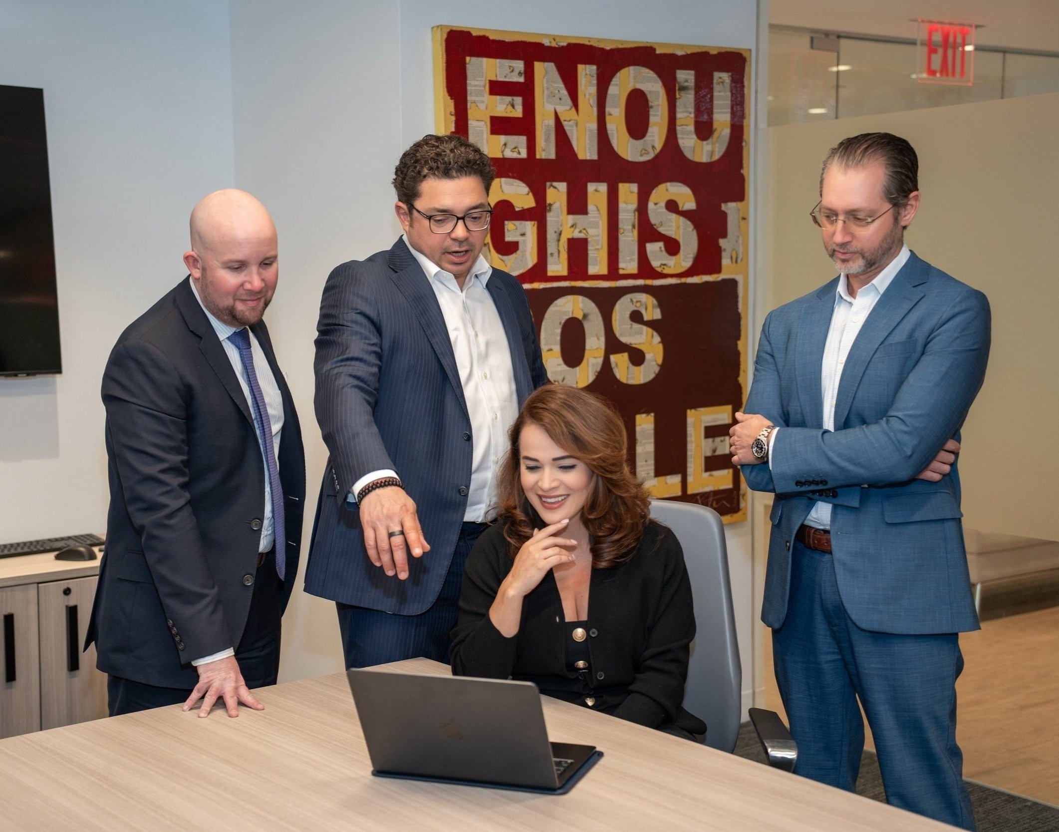 Four men in suits and a woman in a black blazer gathered around a seated woman working on a laptop in an office.