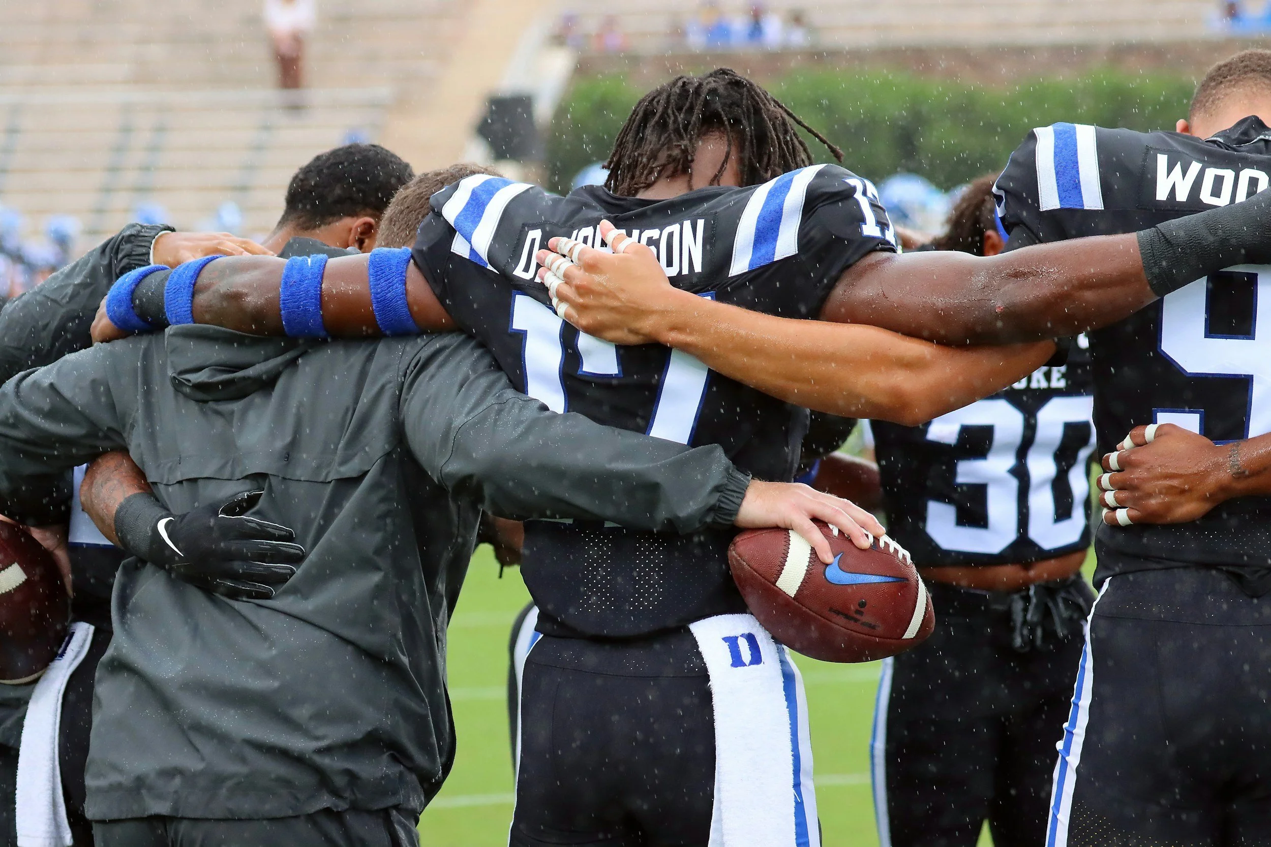 Football players huddled together on the field, some with arms around each other, raining, one holding a football and embracing a coach or staff member.