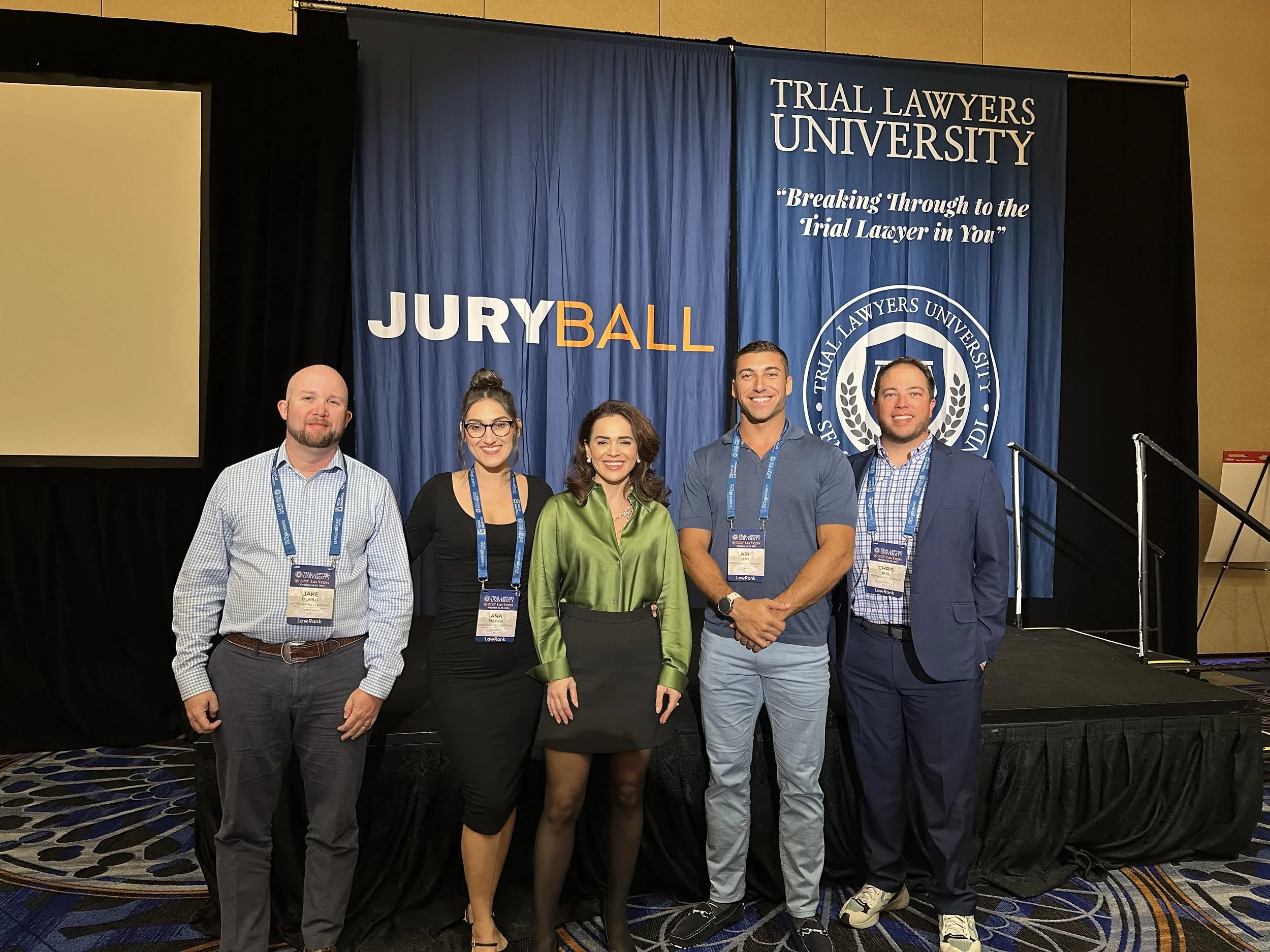 Group of five people standing on stage at a conference, smiling, with a blue backdrop that reads 'TRIAL LAWYERS UNIVERSITY' and 'JURY BALL', with a logo and the slogan 'Breaking Through to the Trial Lawyer in You'.