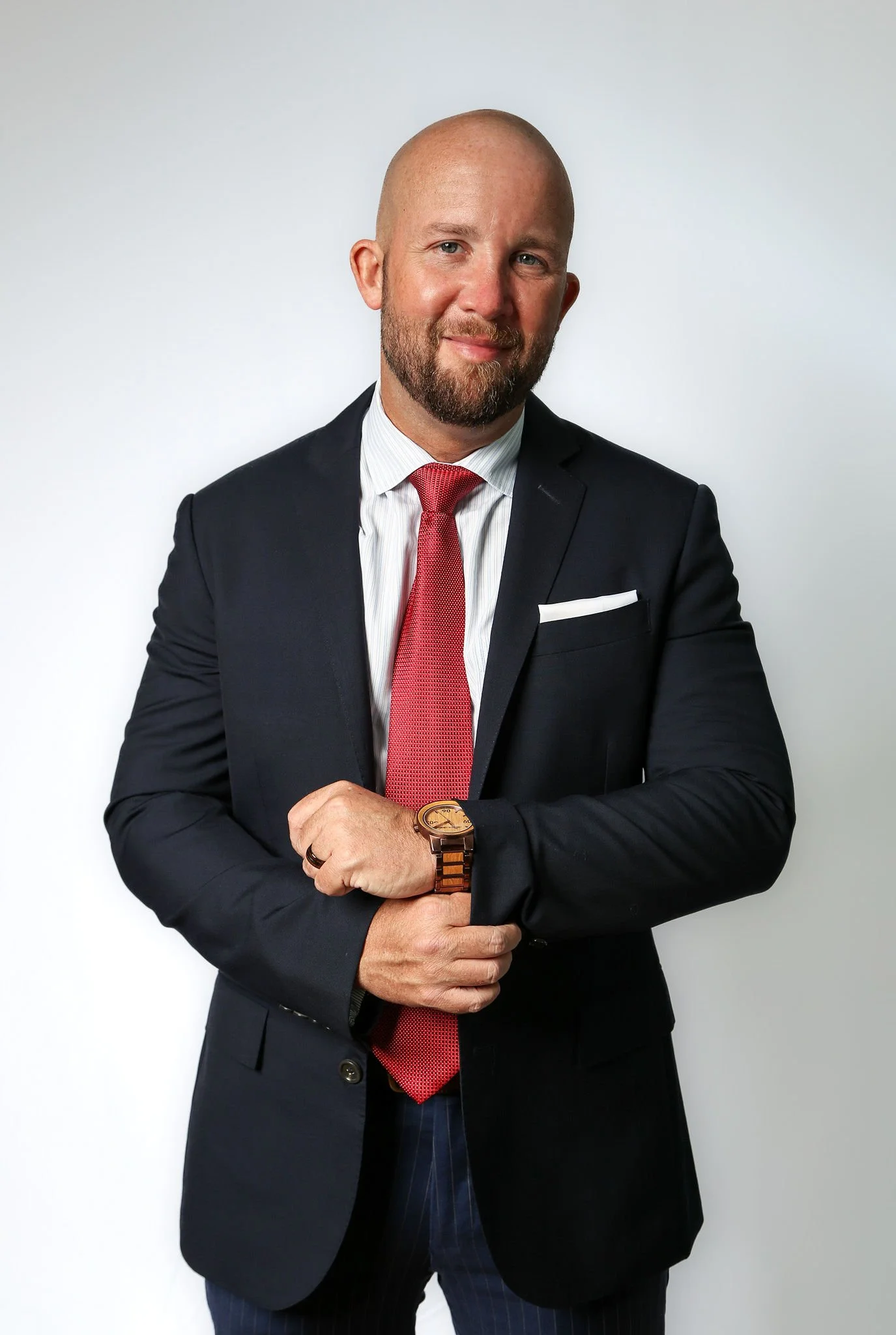 A man in a suit with a red tie, wearing a wristwatch, standing against a plain white background.
