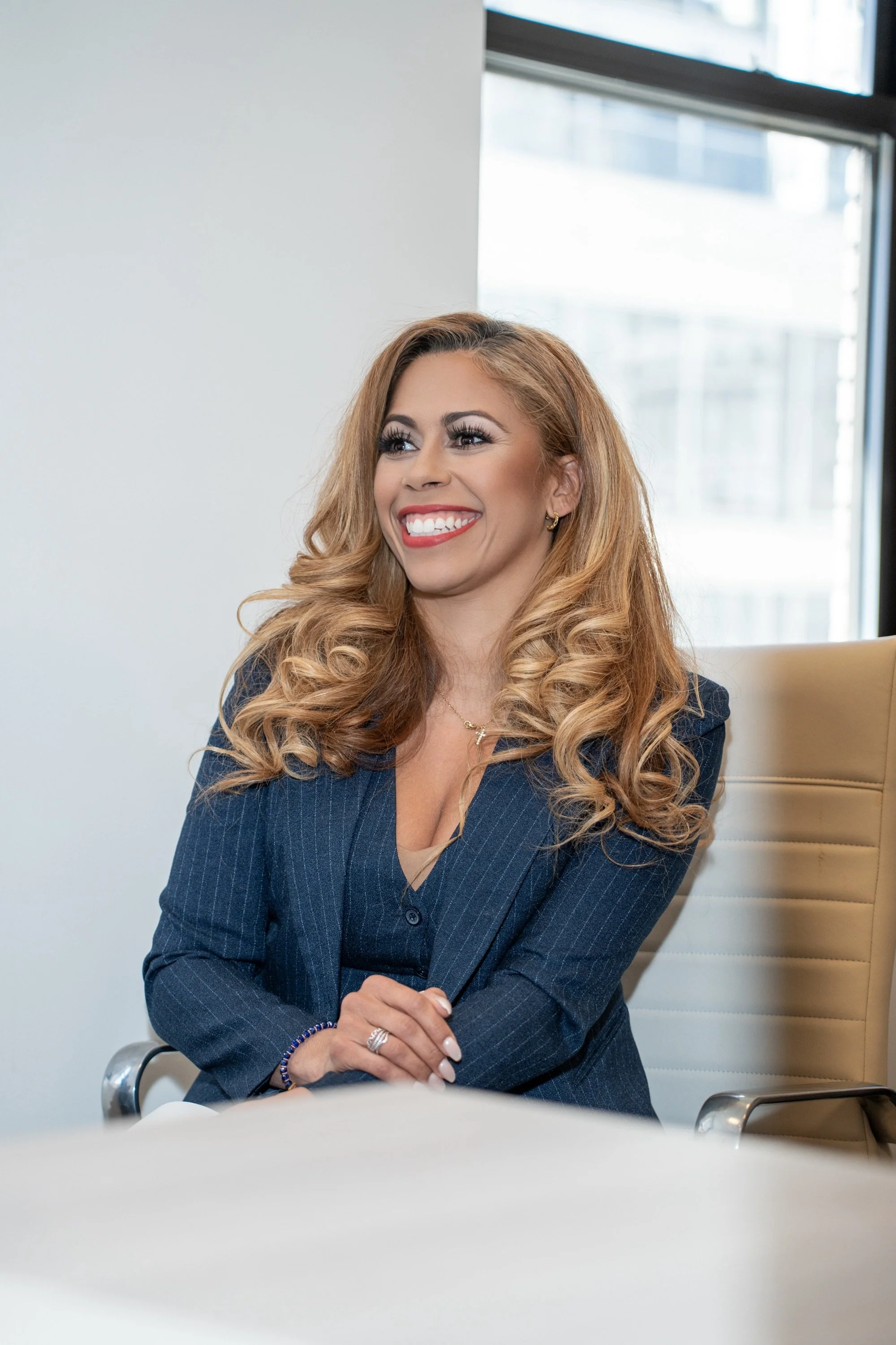 A woman with long, wavy blonde hair, wearing a navy blue pinstripe blazer, smiling and sitting in a bright office with large windows.