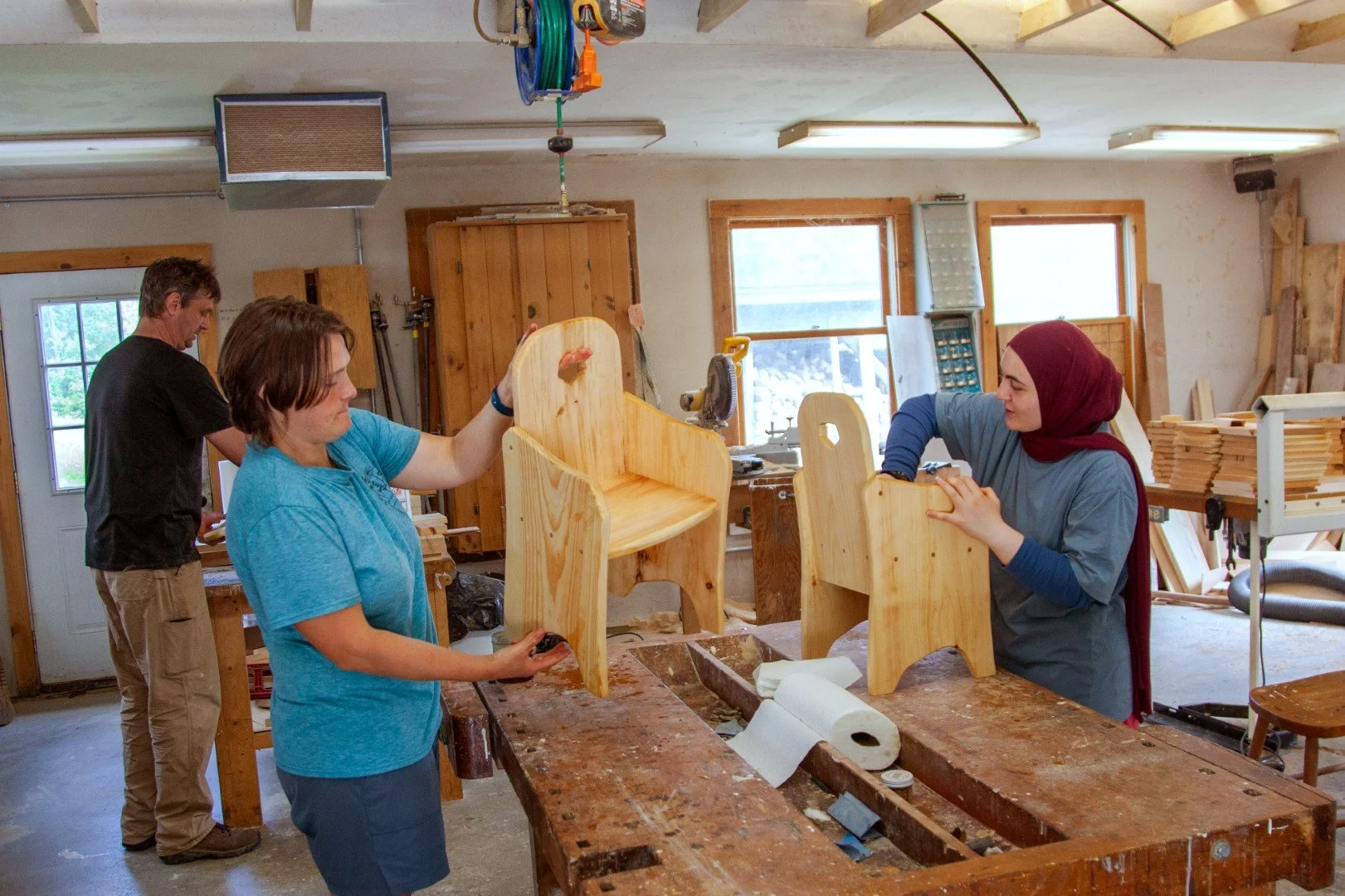 Three people at work in a wood workshop making chairs.