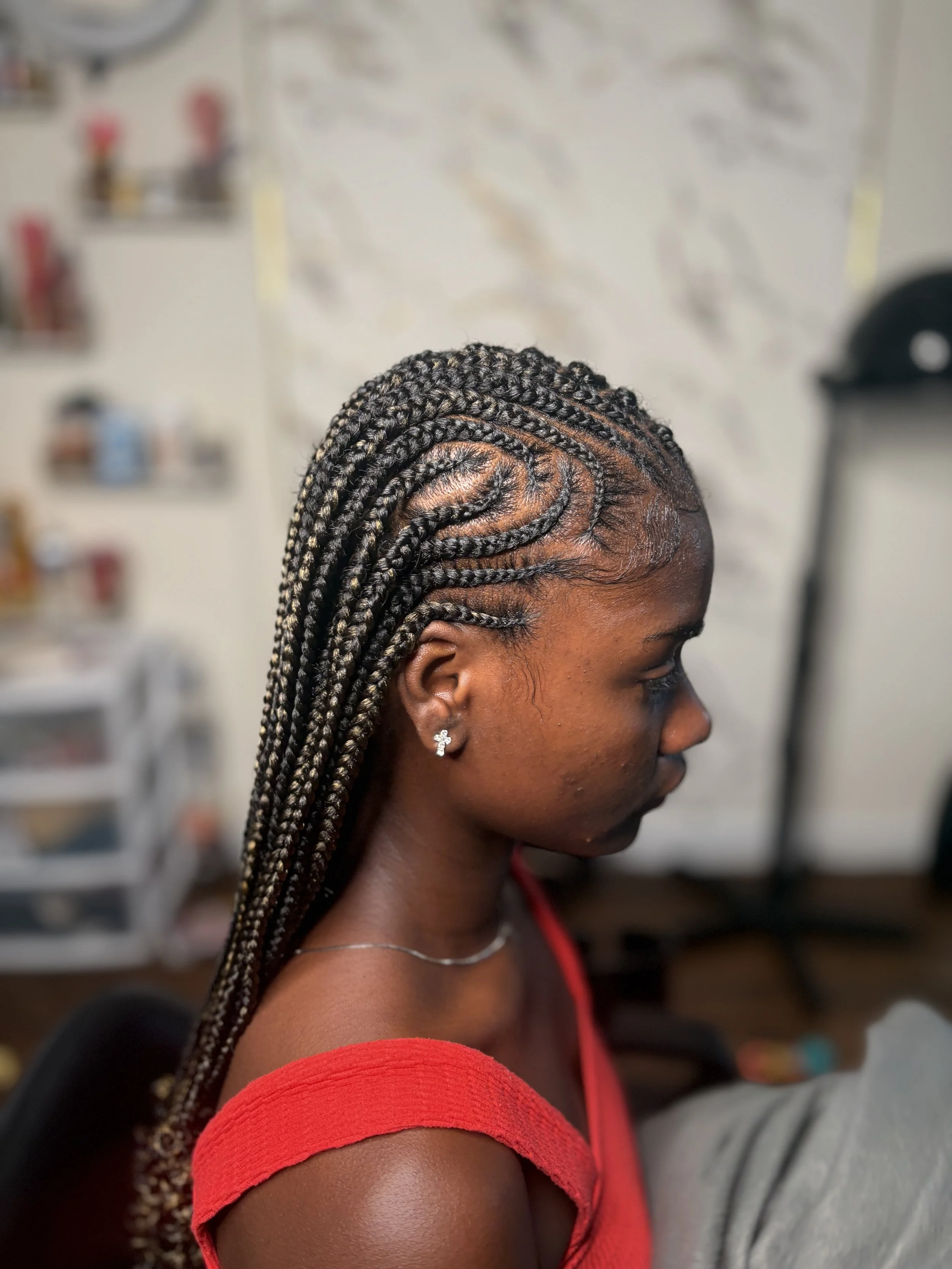 A woman with braided hairstyle sitting indoors, wearing a red top and earrings, with a blurred background.