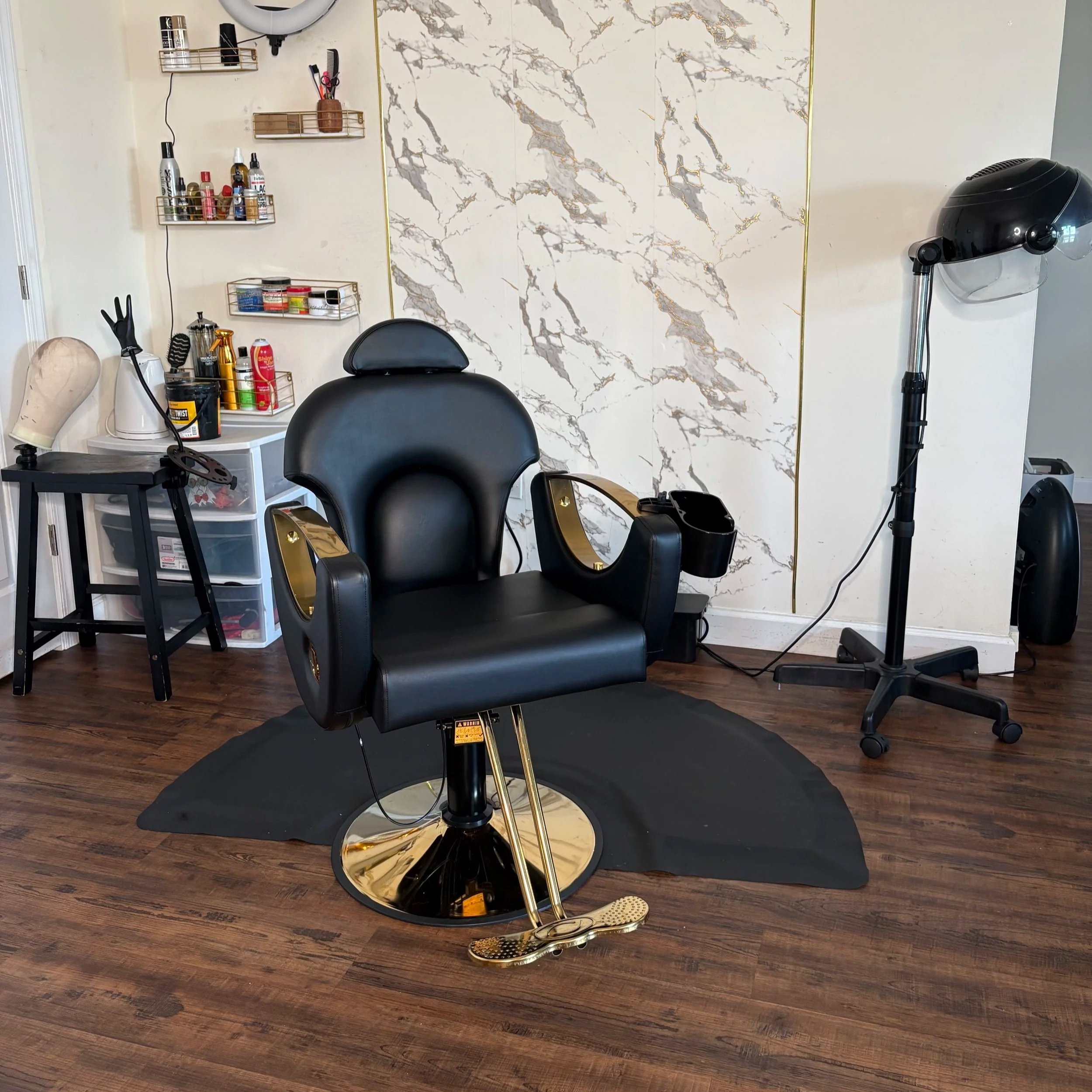 A black barber chair with gold accents in a salon, set on a black mat. To the right, a hair drying or styling hood on a stand. To the left, a small storage unit with tools and products, with shelves above holding various hairstyling supplies. The wall behind features a marble pattern with gold trim.