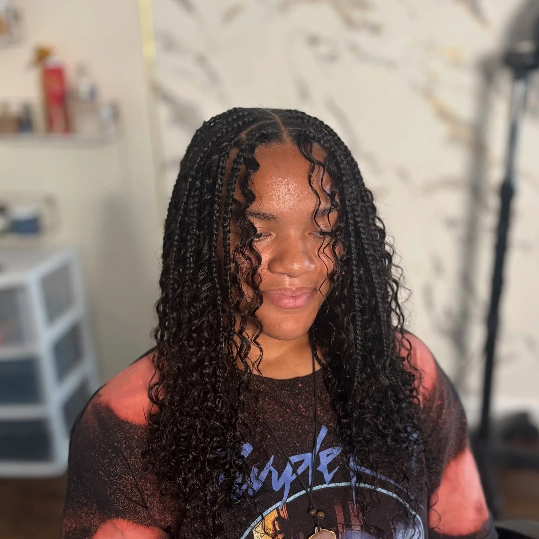 Young woman with long, curly, braided hair and a patterned T-shirt, smiling with her eyes closed in a room with salon or studio equipment in the background.