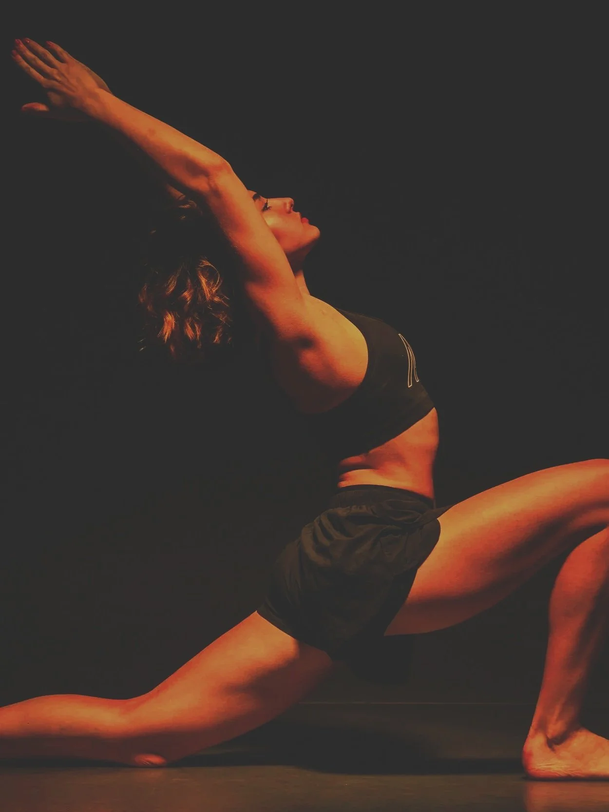 A woman practicing yoga in a lunge pose with her arms raised above her head against a black background.