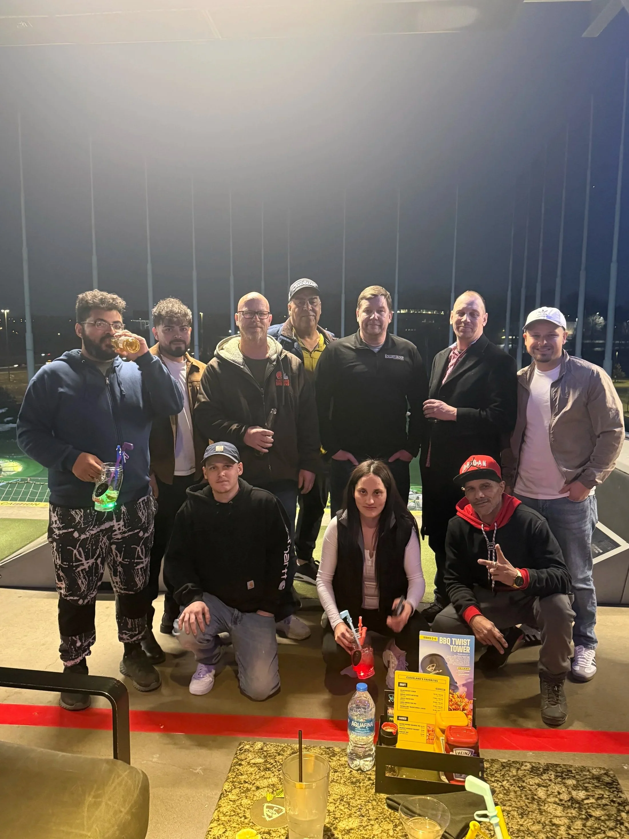 Group of eleven people gathered outdoors at night, some holding drinks, with a golf driving range in the background.