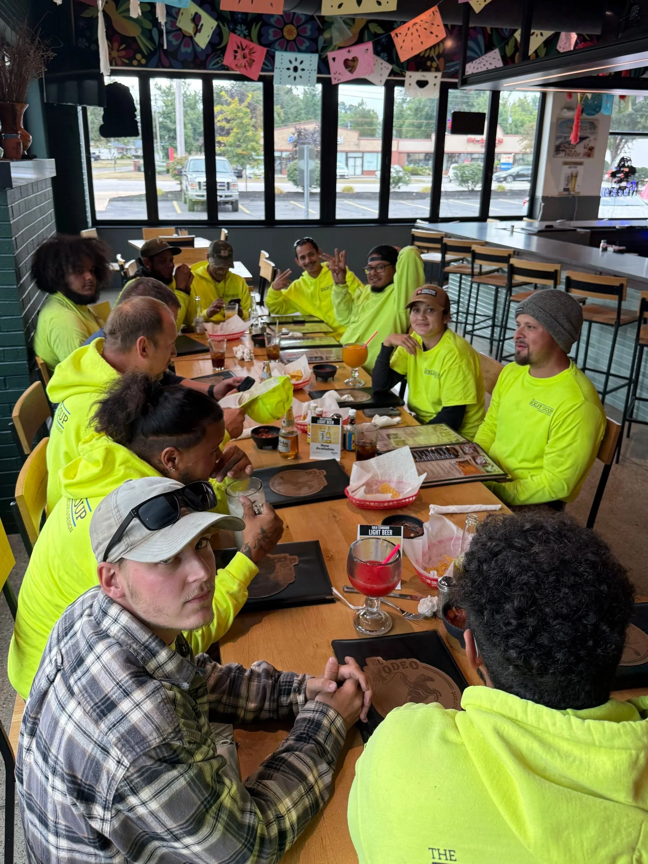 Group of people sitting at a restaurant table wearing matching neon yellow shirts, some holding drinks, with colorful papel picado decorations hanging from the ceiling.
