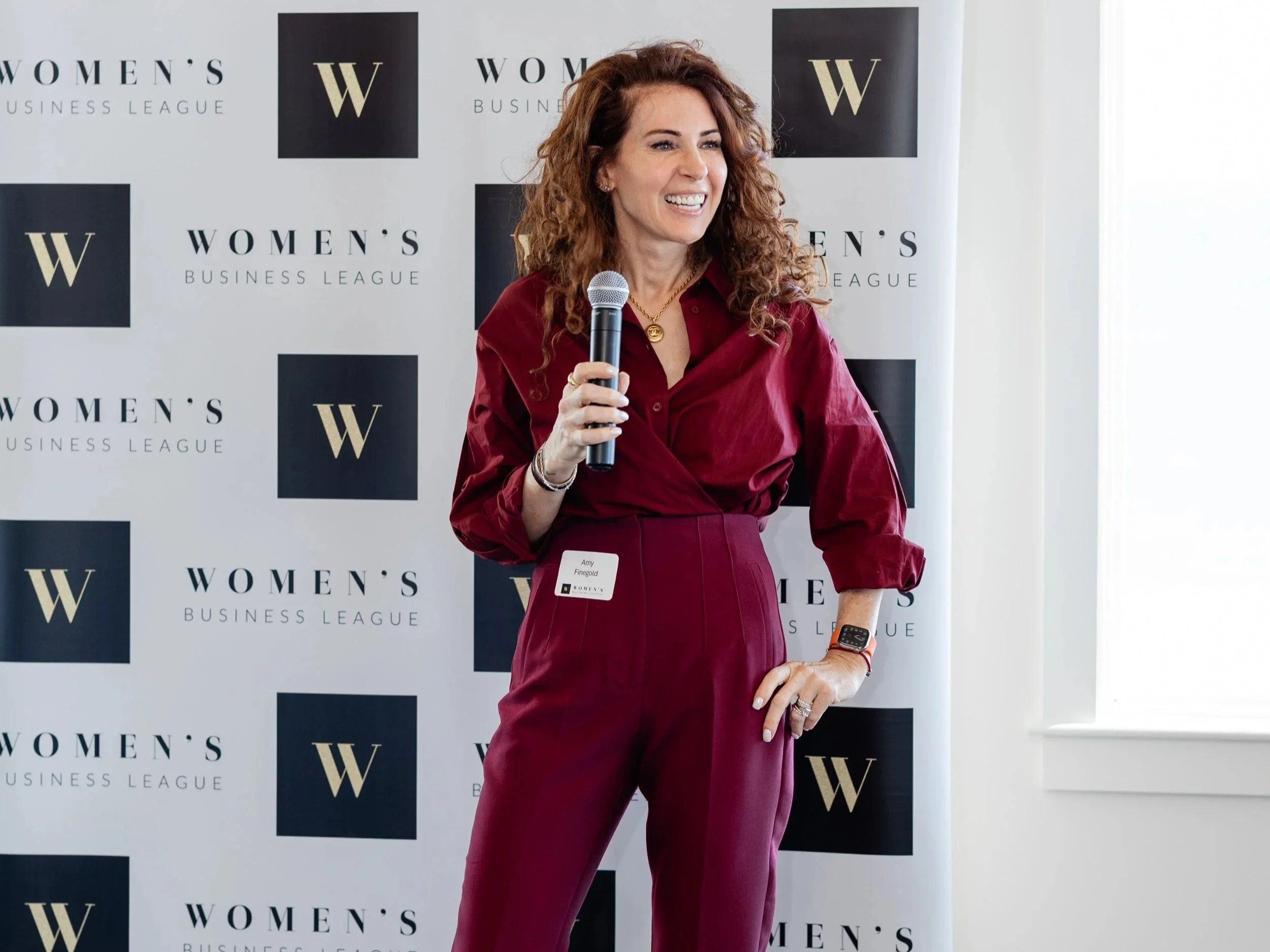 Woman in maroon outfit holding a microphone at Women's Business League event.