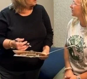 Two women having a conversation in an office. One is standing holding a clipboard and gesturing, the other is sitting and listening.