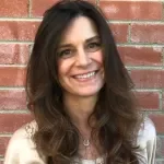 A woman smiling with long dark hair standing against a brick wall.