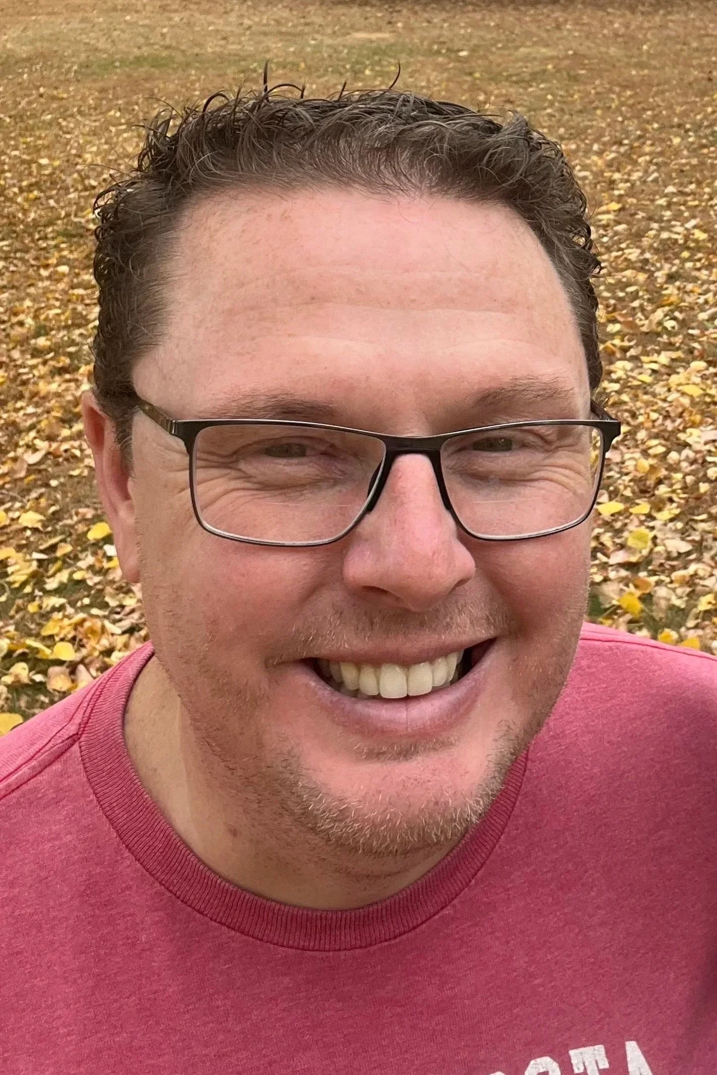 A smiling man with glasses and curly hair outdoors in a field of fallen yellow leaves.