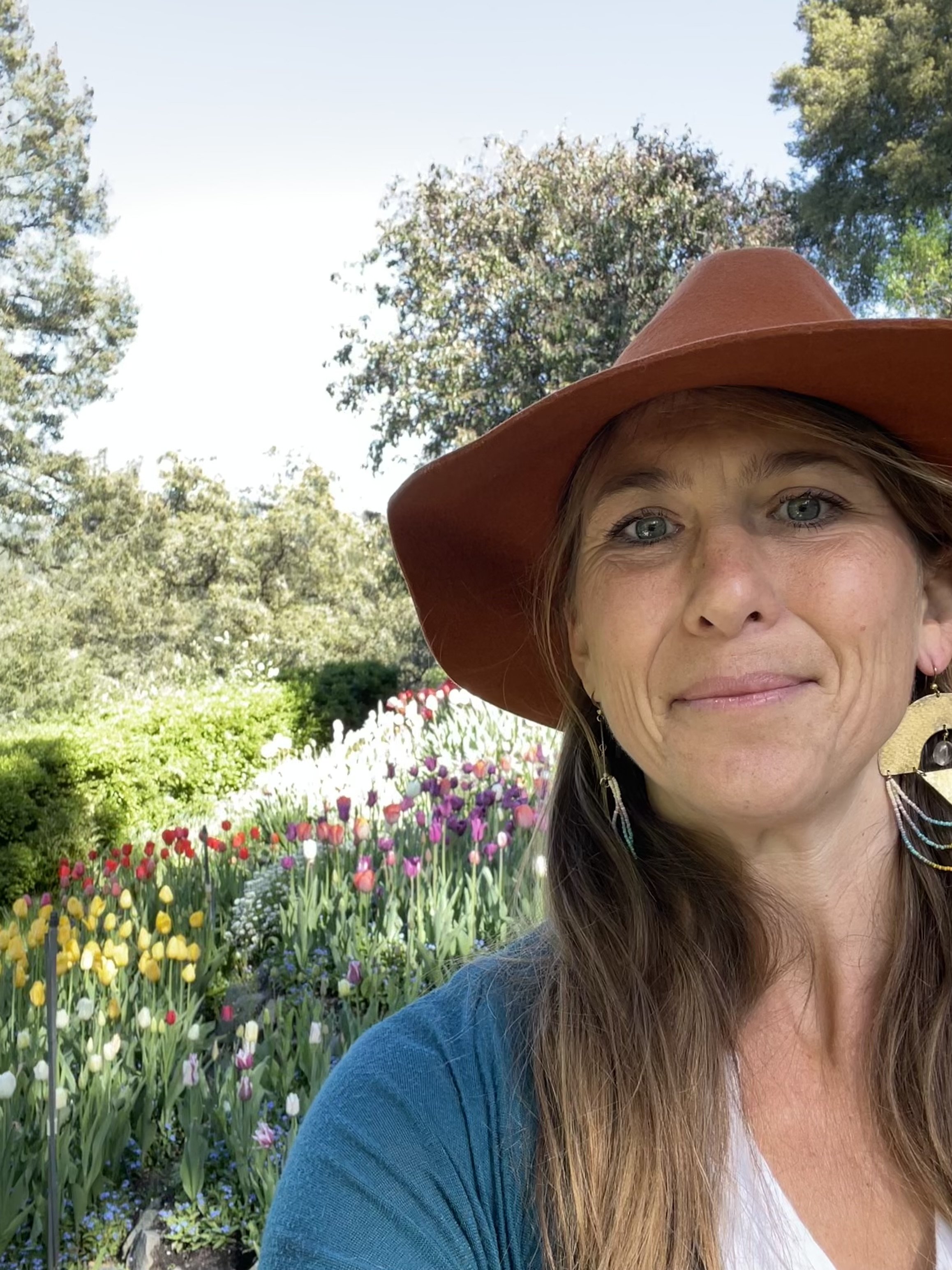 A woman with light skin and brown hair styled with bangs, wearing a black floral patterned jacket with lace details underneath, and red lipstick, standing infront of a tulip garden.
