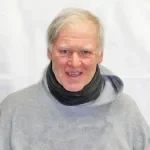 A smiling man with a beard wearing a beige cap and a maroon hoodie with 'Minnesota Trading Co.' on it, standing in front of a blackboard with a clock showing 9:18.