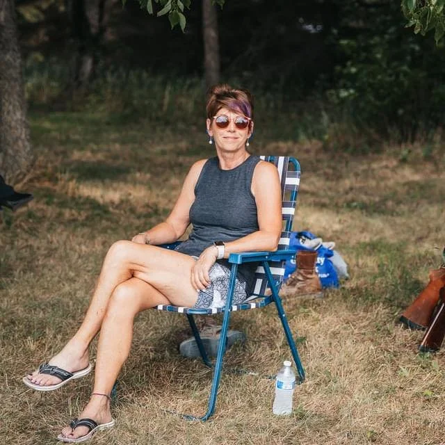 A woman sitting outdoors in a folding chair with trees in the background. She is wearing sunglasses, a sleeveless top, shorts, and flip-flops, with her legs crossed. There is a water bottle on the ground beside her, and some bags and shoes are nearby.