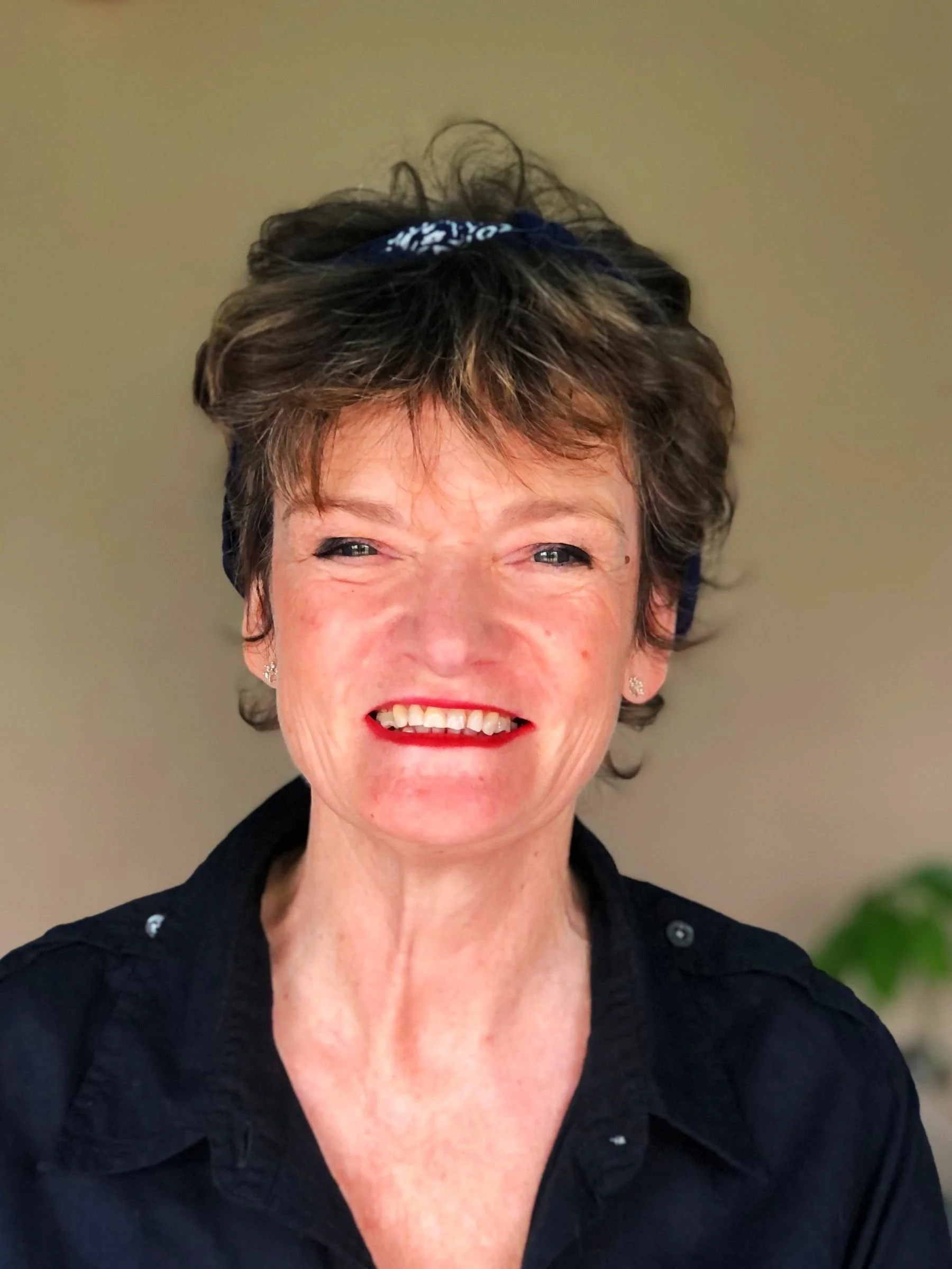 Smiling woman with short, curly brown hair wearing a black shirt and earrings, posing indoors.