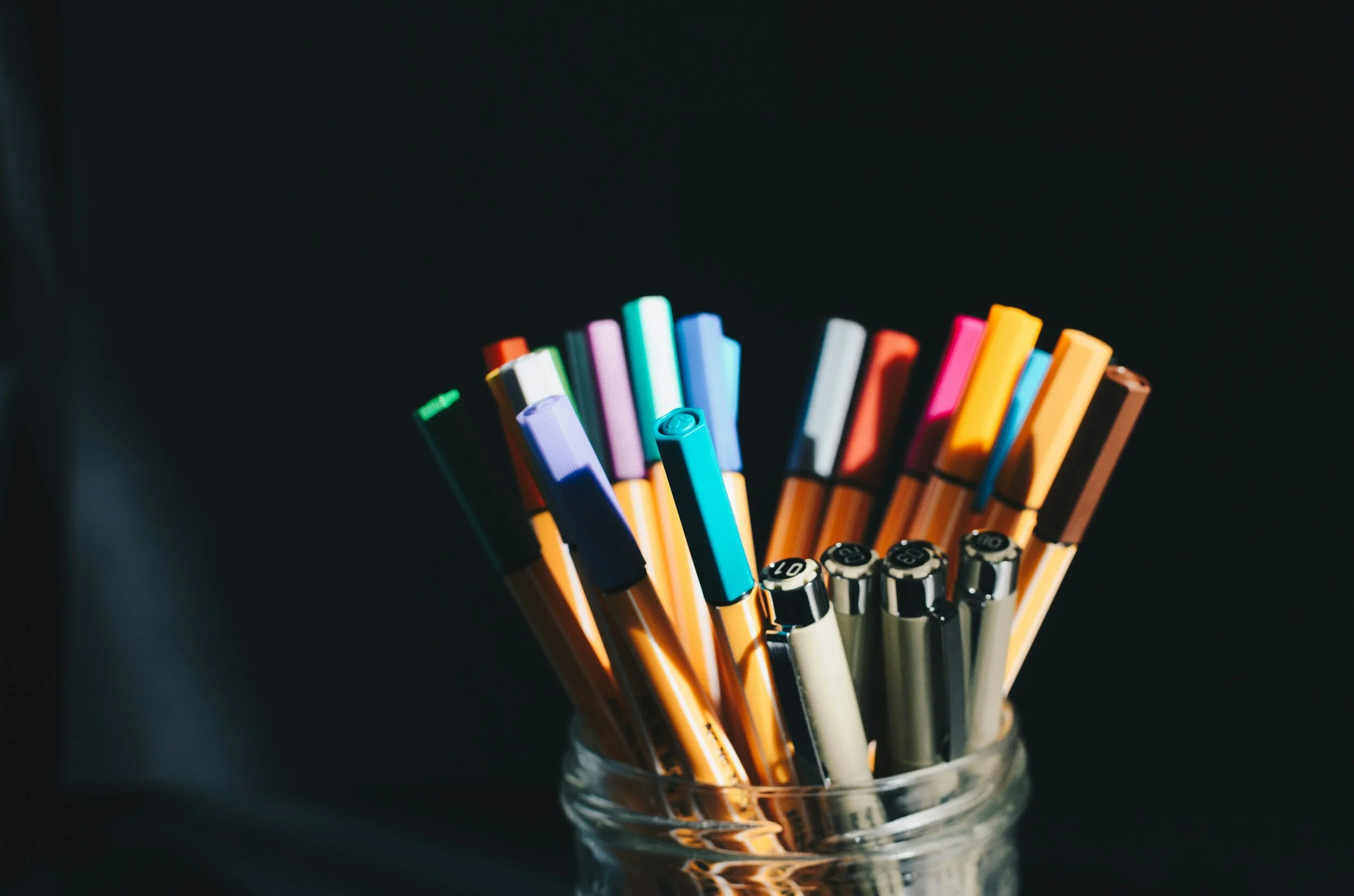 A glass jar filled with colorful markers and highlighters against a dark background.