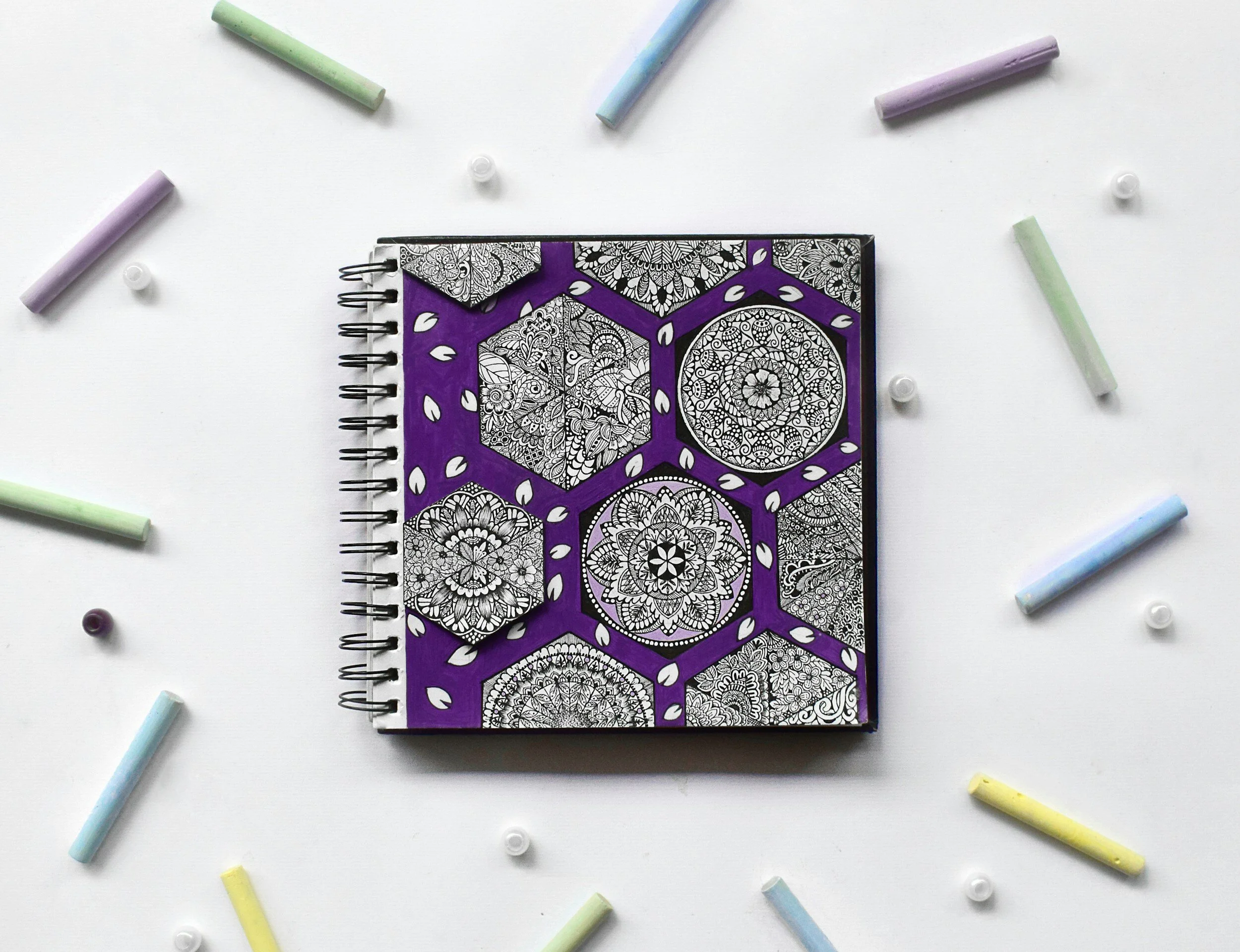 A spiral-bound notebook with intricate black and white mandala and geometric designs on the cover, bordered by purple, surrounded by colorful chalk pieces and small white and black round objects on a white surface.