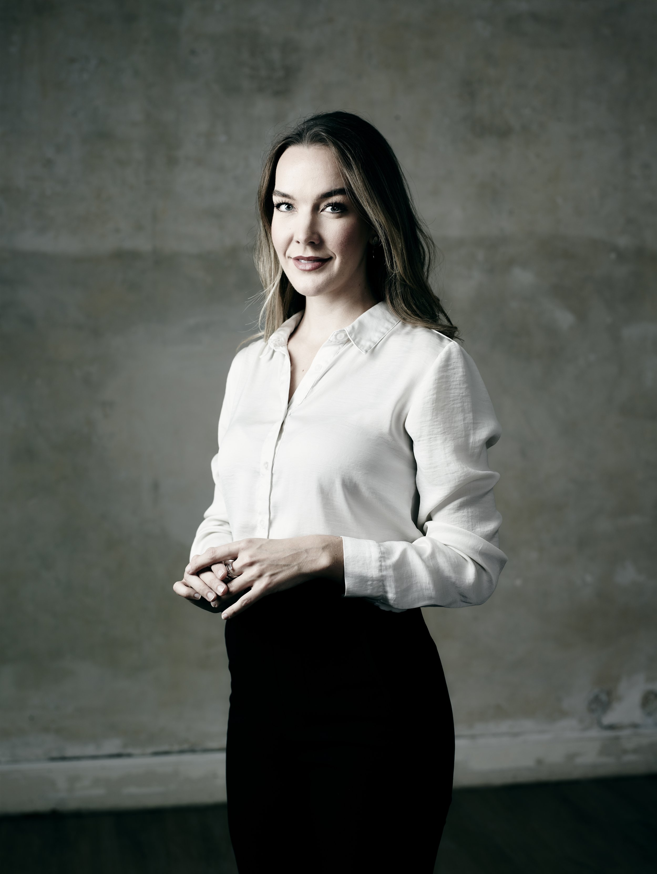 A woman in a white blouse and black skirt standing against a textured wall, looking at the camera with a slight smile.