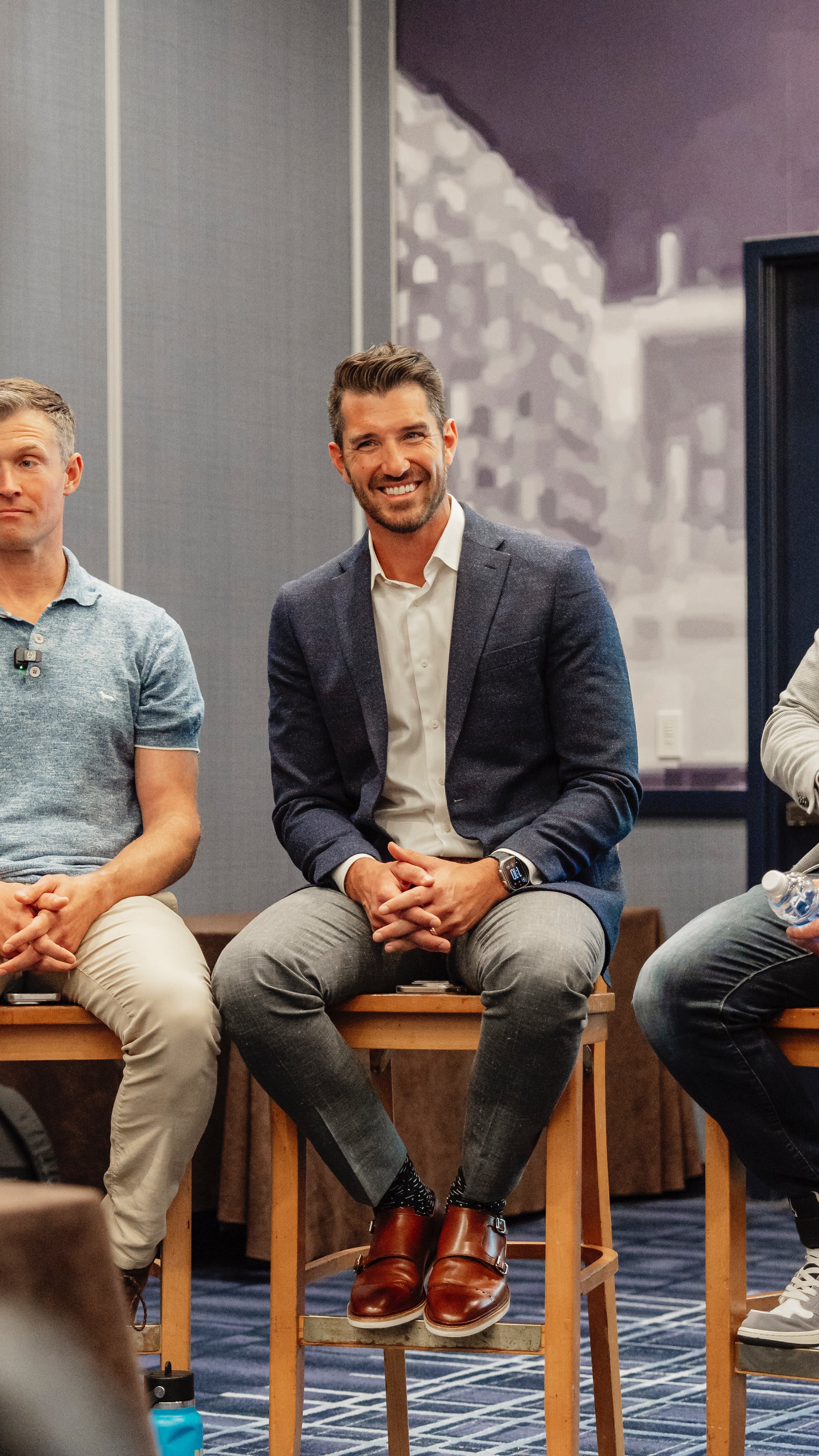 A man in a gray blazer and dress shoes is seated on a wooden stool, smiling and listening during a discussion, in a conference room.