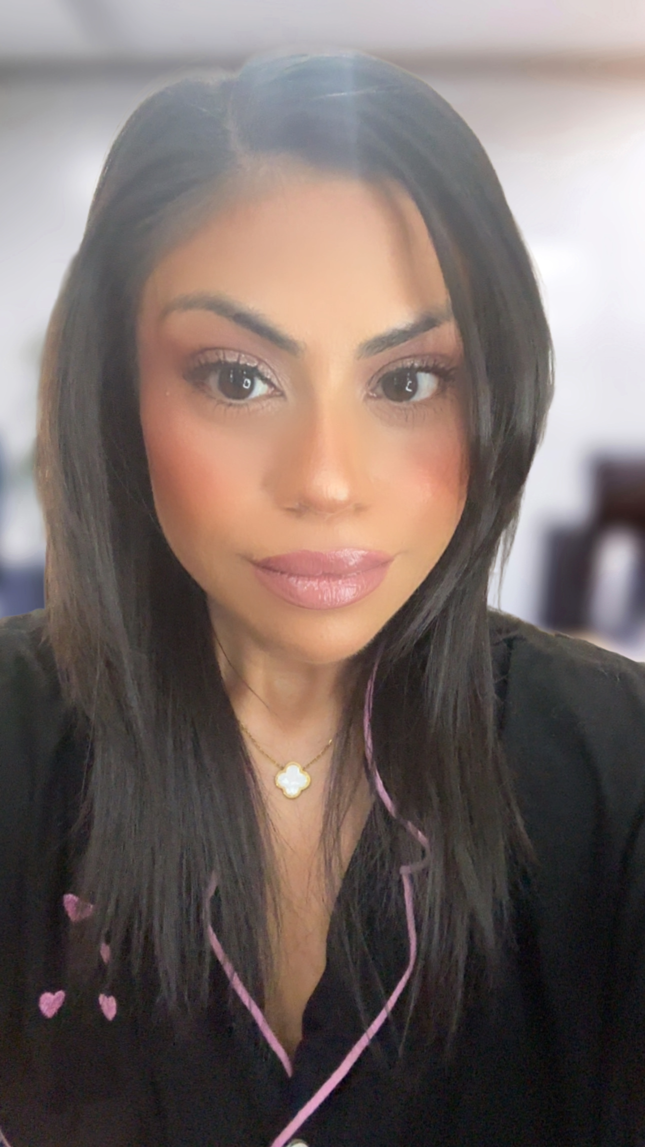 A woman with long wavy dark hair wearing a black blazer, a navy blue top, and a necklace, smiling against a gray background.